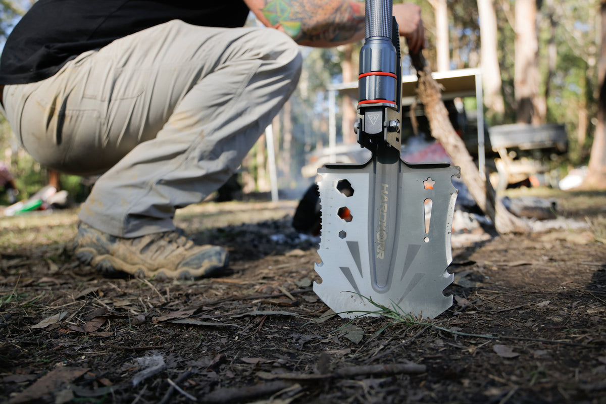 Person kneeling outdoors using a rugged multi-tool shovel in a forest setting, with sunlight filtering through trees and camping gear visible in the background.
