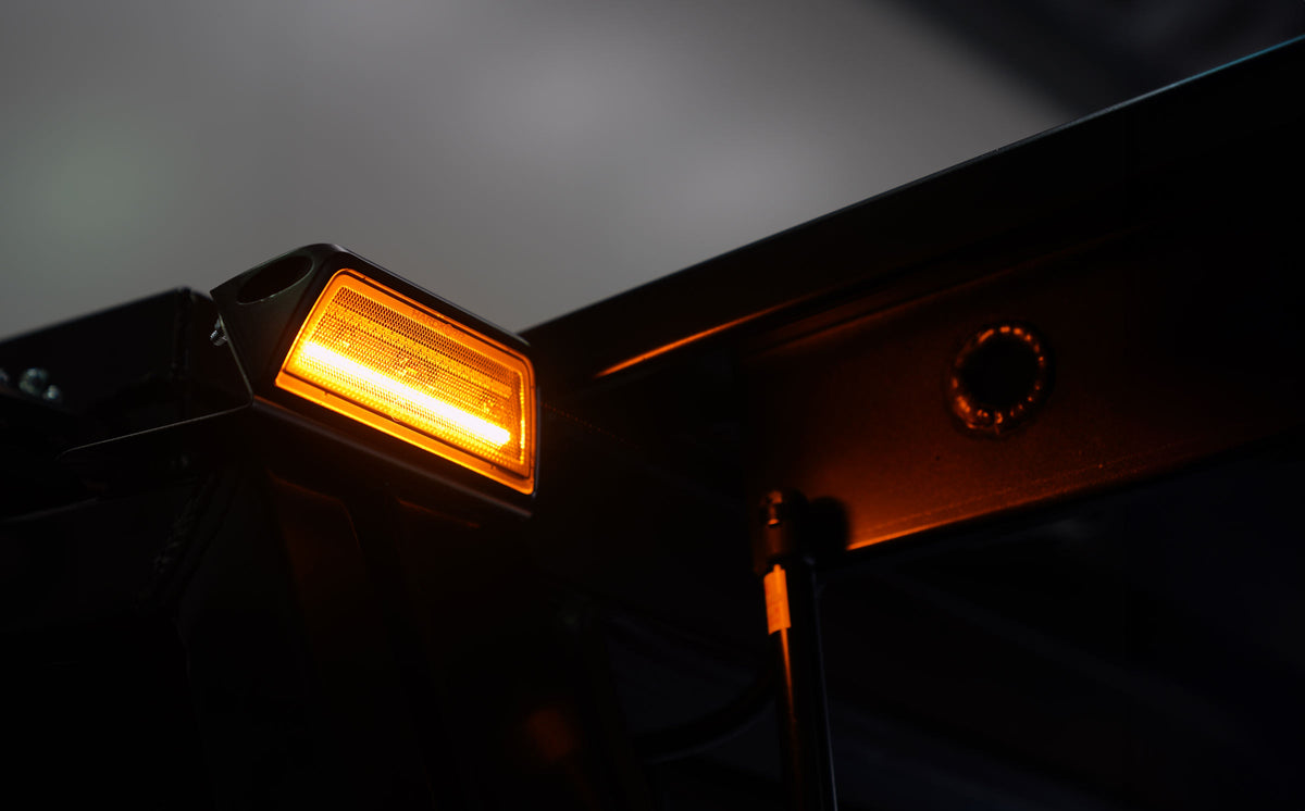 Close-up of an amber LED marker light mounted on a black metal frame, glowing in low light against a dark background, highlighting outdoor vehicle lighting equipment.
