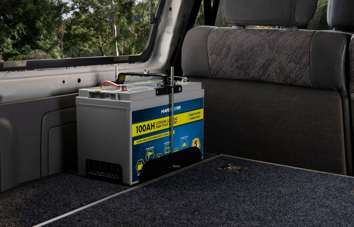 A 100Ah lithium battery securely installed inside the rear cargo area of a vehicle with gray interior and a folded carpeted surface nearby.