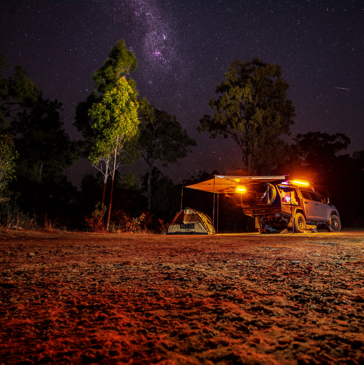 Off-road vehicle set up for camping under a star-filled night sky with an awning extended, illuminated by orange LED camping lights beside a pitched tent near tall trees.