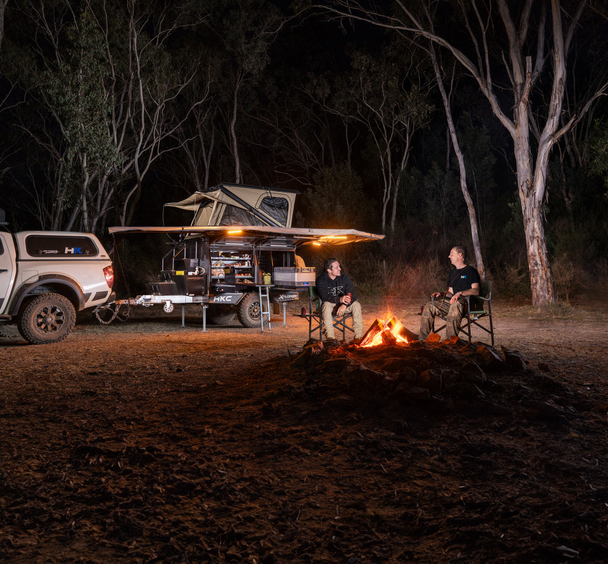 Two people sitting around a campfire at night near a vehicle with an extended canopy and camping gear lit by camping lights in a wooded outdoor setting.