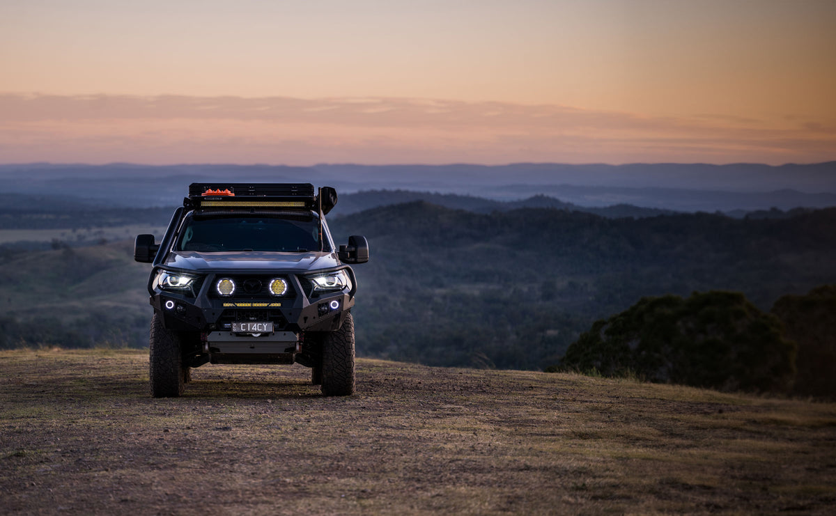Four-wheel-drive vehicle equipped with off-road lights and roof racks parked on a hilltop at sunset, overlooking a vast landscape of rolling hills and trees.