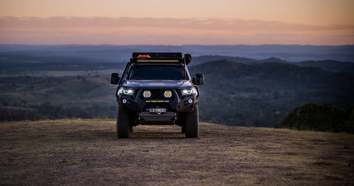 A black 4x4 vehicle equipped with off-road lights and a roof rack, parked on a hilltop at sunset with rolling hills and a colorful sky in the background.