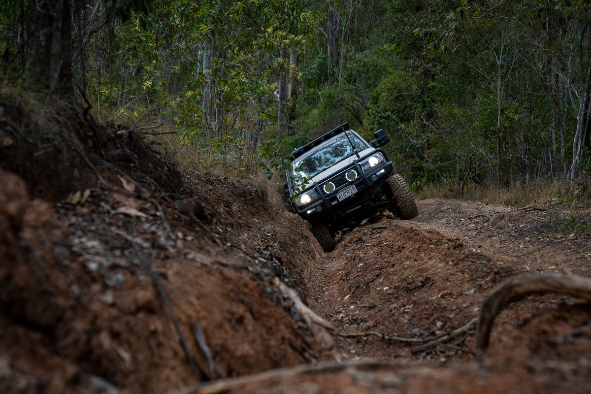 A rugged 4x4 vehicle navigating a deep muddy trail through dense forest terrain under natural daylight.